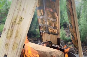 Drei Fischfilets werden zwischen Holzbrettern eingeklemmt und zum Garen im Freien um eine offene Feuerstelle gelegt.